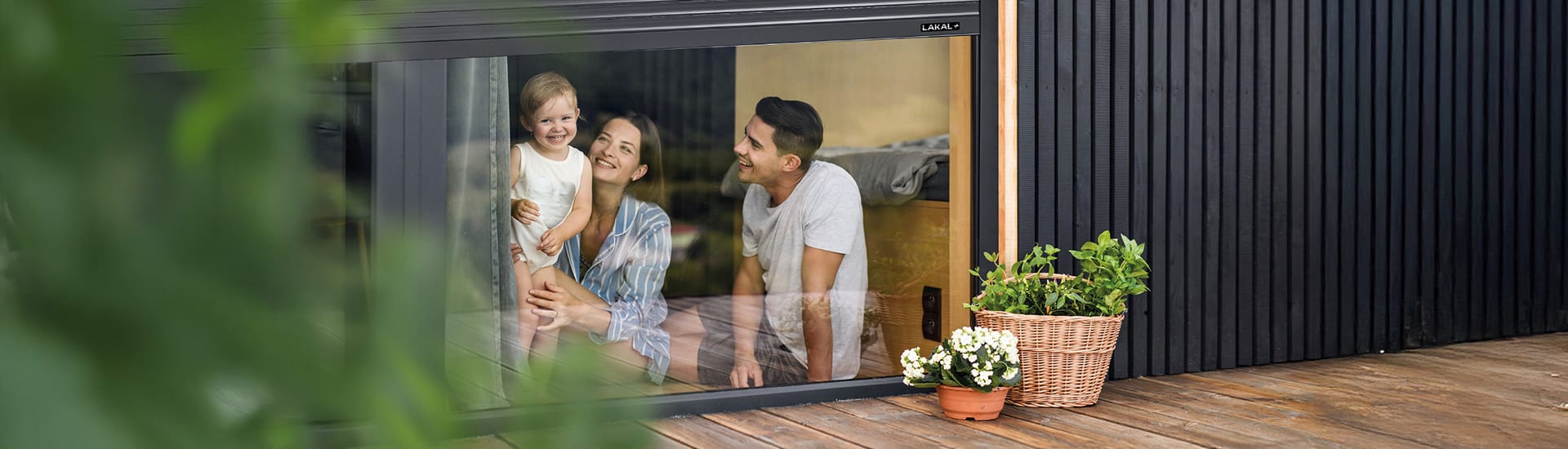 Familie sitzt hinter großen Fensterelementen mit LAKAL Rollladen und genießt ein helles, sicheres Wohnambiente.